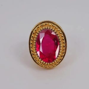 Może przedstawiać: Złoty pierścionek z dużym, owalnym rubinem. Rubin ma intensywny czerwony kolor i jest otoczony teksturowaną, sznurowatą obwódką. Pierścionek jest umieszczony na gładkim, jasnym tle.