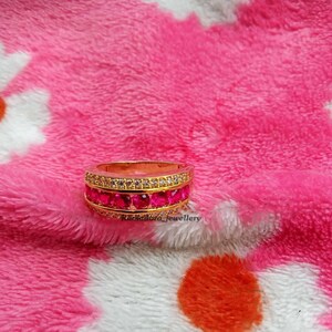 Może przedstawiać: Złoty pierścionek z dwoma rzędami żywych, czerwonych kamieni szlachetnych i górnym rzędem mniejszych, przezroczystych kamieni. Pierścionek jest umieszczony na różowym, teksturowanym tle, podkreślając wzór biżuterii. Pierścionek to biżuteria.