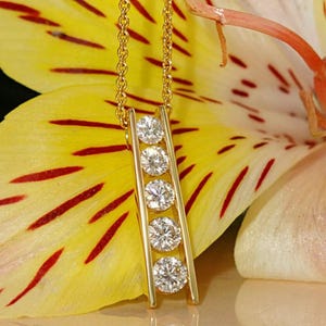 May include: A gold necklace with a vertical bar pendant featuring five round, sparkling diamonds. The chain and pendant are gold-colored, set against a yellow and red floral background. A piece of fine jewelry.