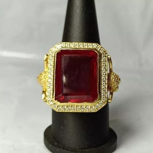 May include: A gold ring with a large, rectangular, deep red gemstone. The gemstone is framed by small, clear stones. The ring has decorative gold detailing on the sides.