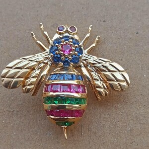 Puede incluir: Broche de abeja dorada con detalles intrincados. El cuerpo de la abeja está adornado con filas de piedras preciosas de colores azul, rosa, verde y rojo. Las alas son texturizadas y los ojos son pequeñas gemas rojas. Un accesorio único y llamativo.