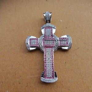 May include: Silver-toned cross pendant with a detailed design. The cross features a combination of white and pink gemstones, with a central circular element. The pendant has a bail for attaching to a chain. The cross is set against a brown background.