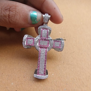May include: A silver cross pendant, adorned with small white and pink gemstones. The cross features a detailed design with a central circular element and arms extending outwards. The pendant is held by a hand. The pendant is likely made of silver.