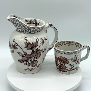 May include: A white ceramic jug and cup with a brown floral pattern. The jug has a handle and a spout. The cup has a handle. Both pieces are decorated with a floral design in shades of brown.