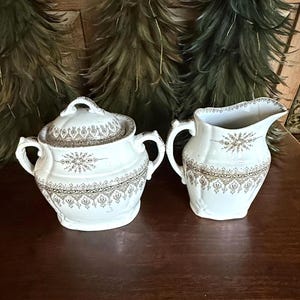 Könnte beinhalten: Weißes Porzellan-Zuckerdose mit Deckel und Sahnekännchen. Beide Teile weisen ein gold-braunes Blumenmuster auf. Die Zuckerdose hat zwei Griffe und einen Deckel mit Griff. Das Sahnekännchen hat einen Griff und einen Ausguss. Das Set steht auf einer dunklen Holzoberfläche.