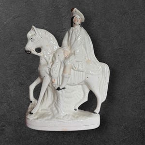 Pode incluir: Uma estatueta de cerâmica branca que retrata uma pessoa a cavalo. O cavaleiro usa chapéu e capa. O cavalo e o cavaleiro apresentam variações sutis de cor. A estatueta é montada em uma base circular.