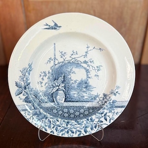 Puede incluir: Un plato de cerámica blanco con un diseño floral y paisajístico azul. El plato presenta una escena central con un jarrón y un paisaje, rodeado de motivos florales y un pájaro volando. El plato se exhibe sobre un soporte transparente.