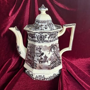 May include: A vintage ceramic teapot with a lid, featuring a black and white landscape design. The teapot has a cream-colored handle, spout, and lid finial. The design includes pagodas, trees, and other architectural elements.