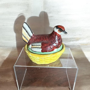 Op de afbeelding: Decoratief keramieken eendenbeeldje met een rood, bruin en wit lichaam, geplaatst op een gele, mandvormige basis. Het beeldje heeft een gedetailleerd ontwerp en staat op een transparante acryl displaystandaard.