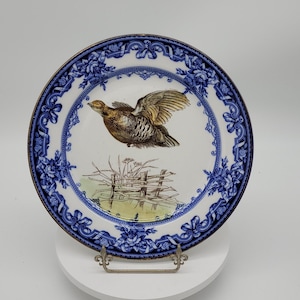 Puede incluir: Un plato de porcelana blanca con un borde azul y dorado. El centro del plato presenta una imagen pintada a mano de un pájaro marrón en vuelo, con una valla y vegetación verde en el fondo.