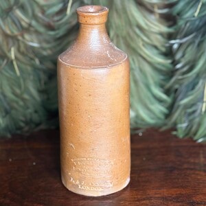 May include: A vintage brown stoneware bottle with a cylindrical body and a narrow neck. The bottle has a slightly textured surface and a label with text. The bottle is likely an antique and is displayed on a wooden surface.
