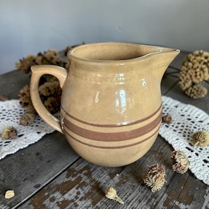 May include: A tan ceramic jug with a handle and spout, featuring two brown horizontal stripes around the body. The jug is set on a rustic wooden surface with dried floral accents and a white lace doily.