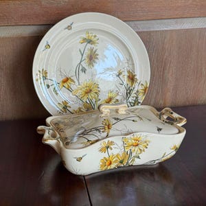 May include: A cream-colored serving dish with a lid and a matching plate. Both pieces are decorated with yellow daisies and green foliage. Gold trim accents the edges and handles. Small bee illustrations are also present.
