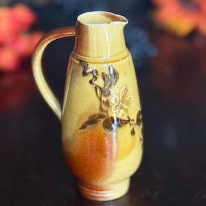 May include: A vintage yellow ceramic pitcher with a brown handle and floral design. The pitcher has a rounded body and a spout for pouring. The floral design features brown flowers and leaves. The pitcher is likely used for serving beverages.