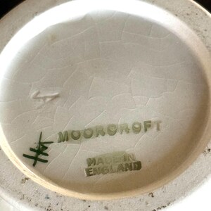 May include: Close-up of the base of a ceramic vase. The text "MOORCROFT" and "MADE IN ENGLAND" are printed in green. The base is off-white with a cracked glaze.