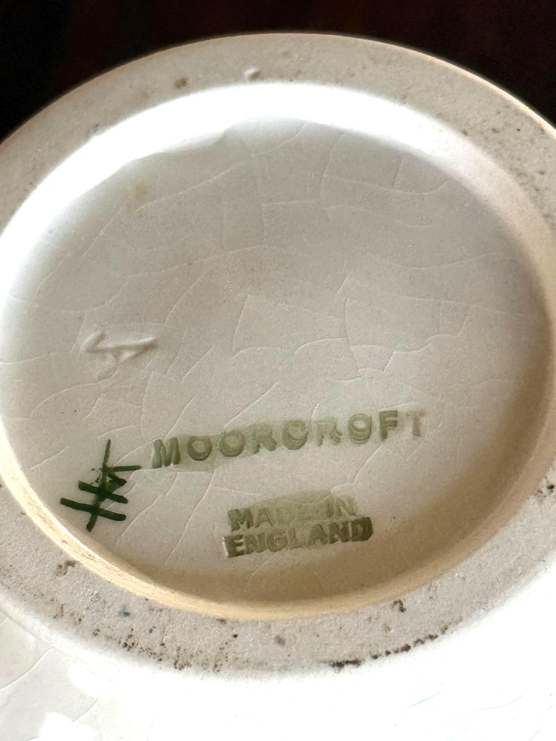 May include: Close-up of the base of a ceramic vase. The text "MOORCROFT" and "MADE IN ENGLAND" are printed in green. The base is off-white with a cracked glaze.