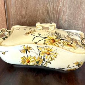 May include: Cream-colored ceramic serving dish with a lid, adorned with yellow daisy floral patterns and gold accents. The dish has a rectangular shape with curved edges and handles, perfect for serving food.