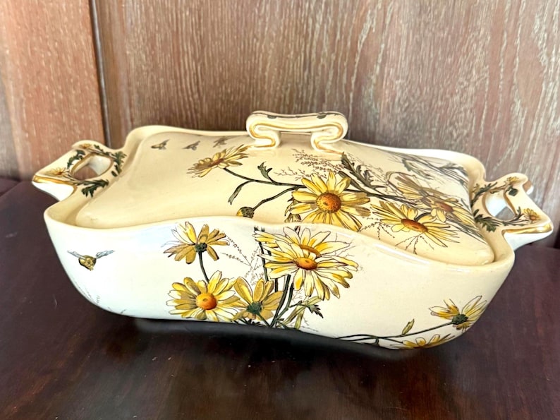 May include: Cream-colored ceramic serving dish with a lid, adorned with yellow daisy floral patterns and gold accents. The dish has a rectangular shape with curved edges and handles, perfect for serving food.