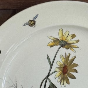 May include: Off-white ceramic plate with a floral design featuring yellow daisies and green stems. Two small bees with blue wings are also depicted. The plate has a slightly aged appearance.