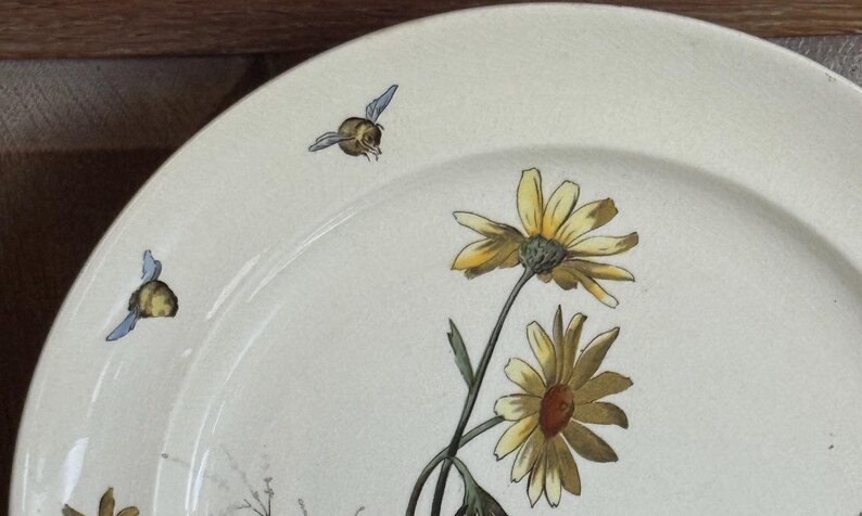 May include: Off-white ceramic plate with a floral design featuring yellow daisies and green stems. Two small bees with blue wings are also depicted. The plate has a slightly aged appearance.