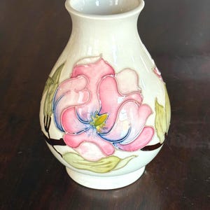 May include: Cream-colored ceramic vase with a floral design. The vase features a large pink and blue flower with green leaves. The vase has a narrow neck and a round base. The floral design is hand-painted.