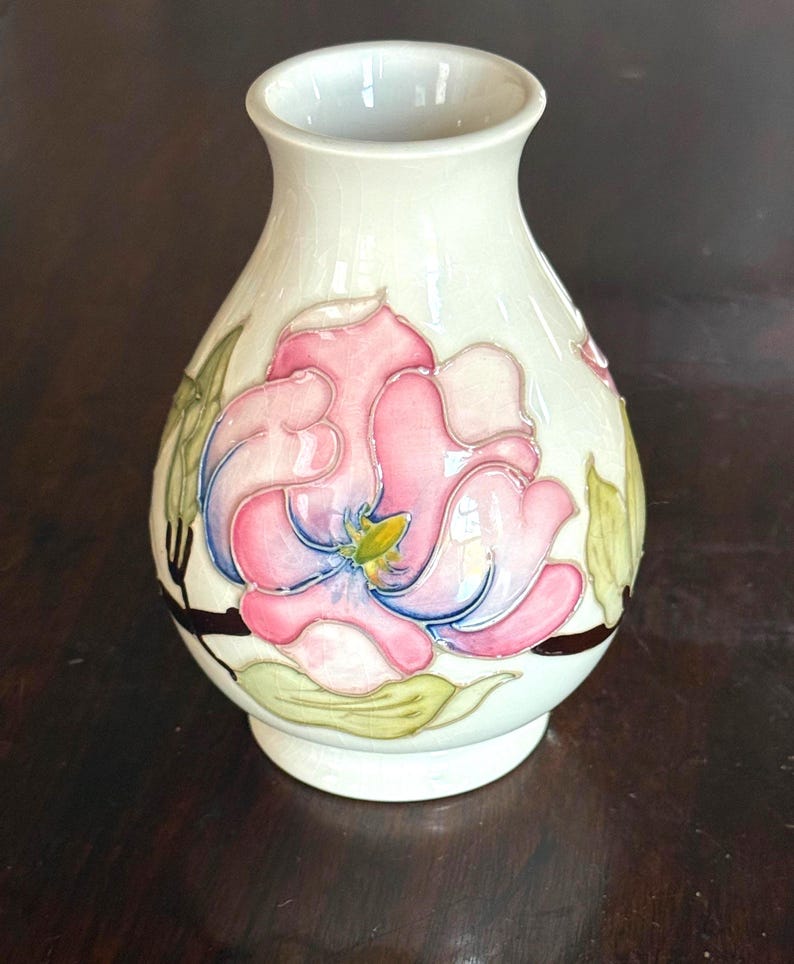 May include: Cream-colored ceramic vase with a floral design. The vase features a large pink and blue flower with green leaves. The vase has a narrow neck and a round base. The floral design is hand-painted.