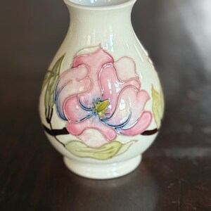 May include: A cream-colored ceramic vase with a floral design. The vase features a large pink and blue flower with green leaves and a yellow center. The vase has a rounded body and a slightly flared opening.
