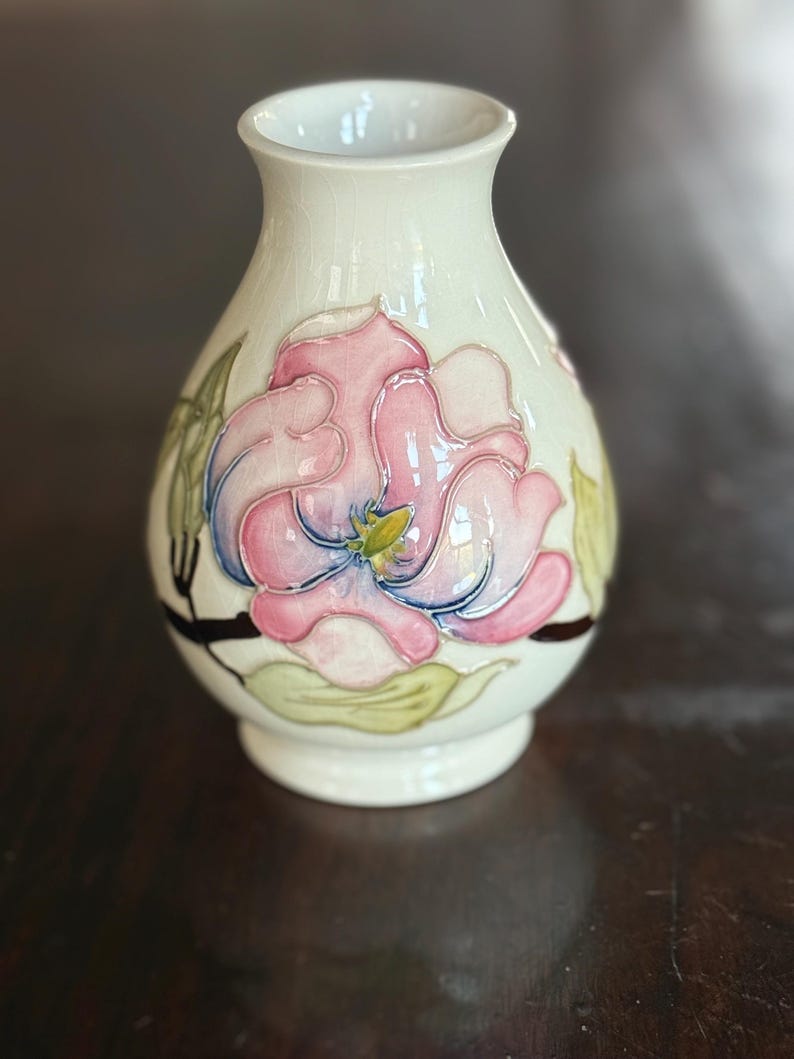 May include: A cream-colored ceramic vase with a floral design. The vase features a large pink and blue flower with green leaves and a yellow center. The vase has a rounded body and a slightly flared opening.