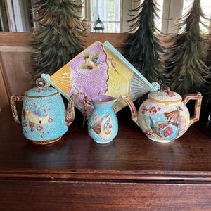 Może przedstawiać: Kolekcja zabytkowych ceramicznych filiżanek i imbryków do herbaty z ozdobnymi uchwytami i pokrywkami. Zestaw zawiera talerz w kształcie wachlarza i mały dzbanek, wszystkie zdobione motywami kwiatowymi i ptasimi w pastelowych kolorach. Przedmioty są ułożone na ciemnej drewnianej powierzchni.