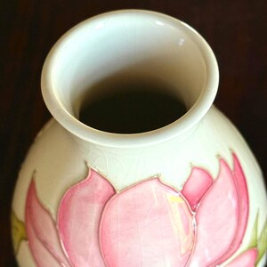 May include: Cream-colored ceramic vase with a wide opening and a rounded body. The vase is decorated with a pink lotus flower design, with green and blue accents. The vase has a glossy finish.