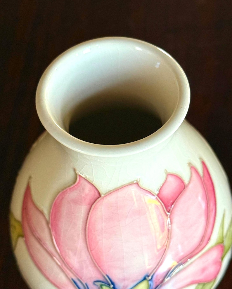May include: Cream-colored ceramic vase with a wide opening and a rounded body. The vase is decorated with a pink lotus flower design, with green and blue accents. The vase has a glossy finish.