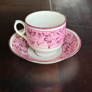 Puede incluir: Un juego de taza y platillo vintage. La taza y el platillo son blancos con un diseño floral rosa y un borde rosa. La taza tiene un asa pequeña. El juego está colocado sobre una superficie de madera marrón oscuro.
