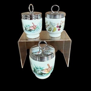 May include: Three decorative egg cups with silver lids and handles. The porcelain cups feature hand-painted designs of birds, berries, and foliage. The cups are displayed on a clear, tiered stand against a black background.