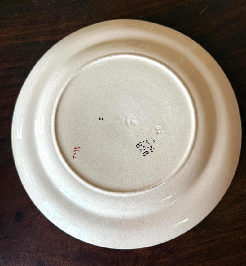 May include: Off-white ceramic plate with a wide rim. The underside features stamped text including "R No. 826" and "7496". The plate has a slightly aged appearance, suggesting it is vintage or antique.