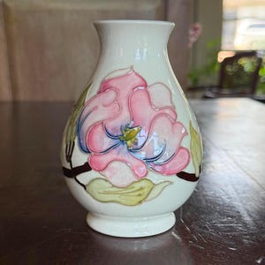May include: A small, off-white ceramic vase with a hand-painted floral design. The pink and blue flower is the focal point, with green leaves and brown stems. The vase has a rounded body and a narrow neck.