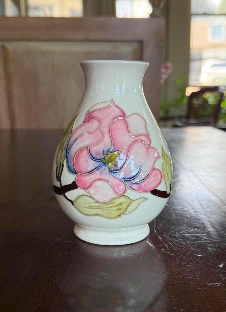 May include: A small, off-white ceramic vase with a hand-painted floral design. The pink and blue flower is the focal point, with green leaves and brown stems. The vase has a rounded body and a narrow neck.