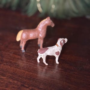 Puede incluir: Un caballo de juguete marrón con crin y cola beige está detrás de un perro de juguete blanco y marrón manchado. El perro tiene un collar marrón. Ambos juguetes están sobre una superficie de madera oscura.