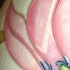 May include: Close-up of a ceramic piece featuring a stylized pink flower with detailed veining. The petals are a soft pink with darker pink outlines and blue accents. The background is a light cream color with a cracked glaze effect.