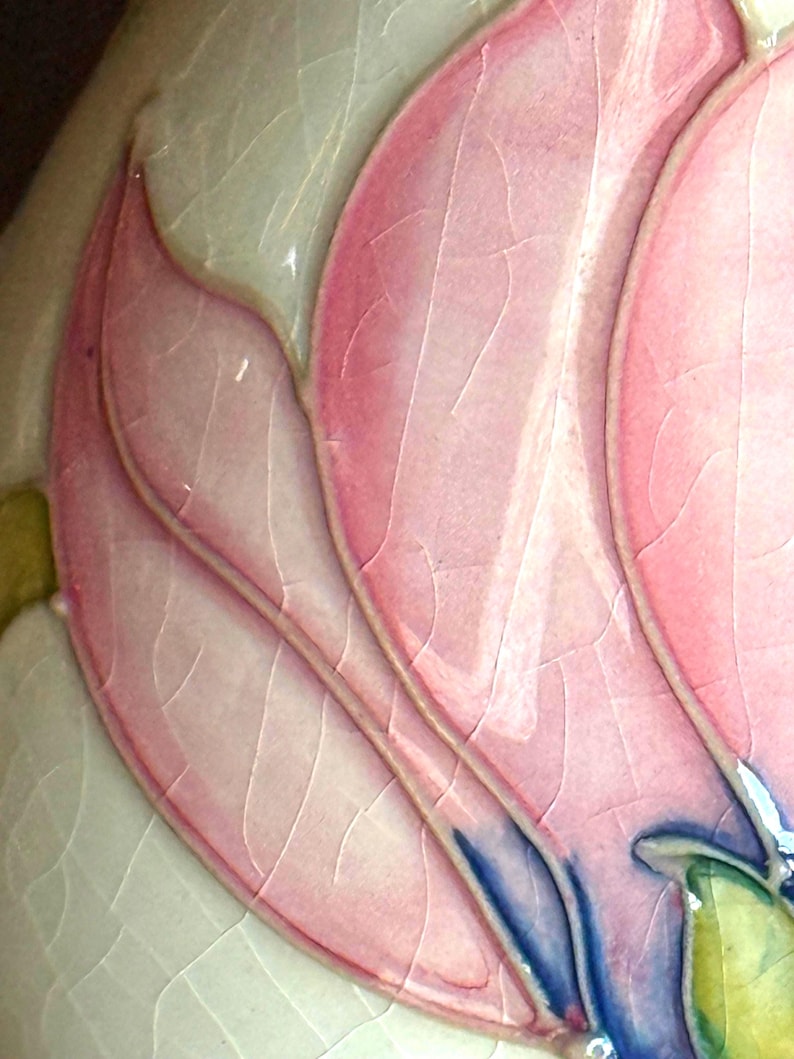 May include: Close-up of a ceramic piece featuring a stylized pink flower with detailed veining. The petals are a soft pink with darker pink outlines and blue accents. The background is a light cream color with a cracked glaze effect.