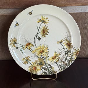 May include: A cream-colored ceramic plate featuring a floral design of yellow daisies and green stems. Two small bees are also depicted. The plate is displayed on a gold-colored metal stand, showcasing a vintage aesthetic.