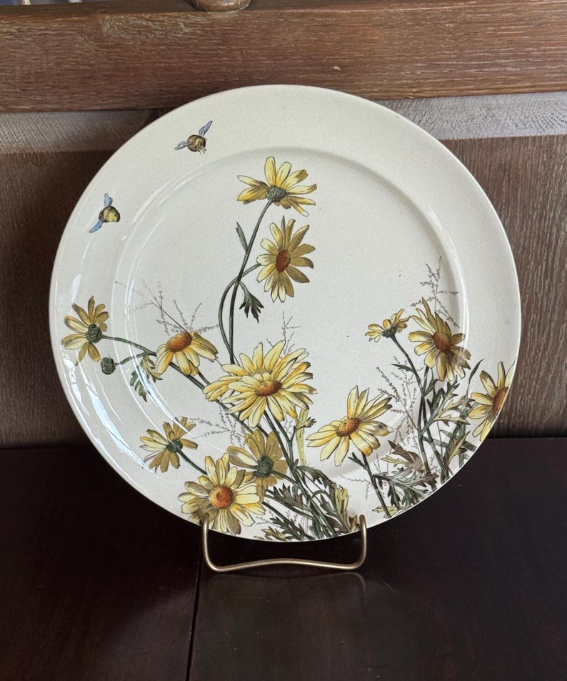 May include: A cream-colored ceramic plate featuring a floral design of yellow daisies and green stems. Two small bees are also depicted. The plate is displayed on a gold-colored metal stand, showcasing a vintage aesthetic.