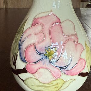 May include: Cream-colored ceramic vase with a large, raised floral design. The flower features pink petals with blue and yellow accents, surrounded by green leaves and brown stems. The vase has a crackled glaze.