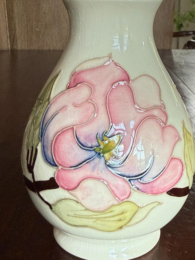 May include: Cream-colored ceramic vase with a large, raised floral design. The flower features pink petals with blue and yellow accents, surrounded by green leaves and brown stems. The vase has a crackled glaze.