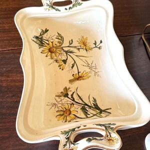 May include: Cream-colored rectangular serving tray with a scalloped edge and two handles. The tray is decorated with yellow daisies and green leaves. The handles are adorned with floral accents. A decorative piece for serving.