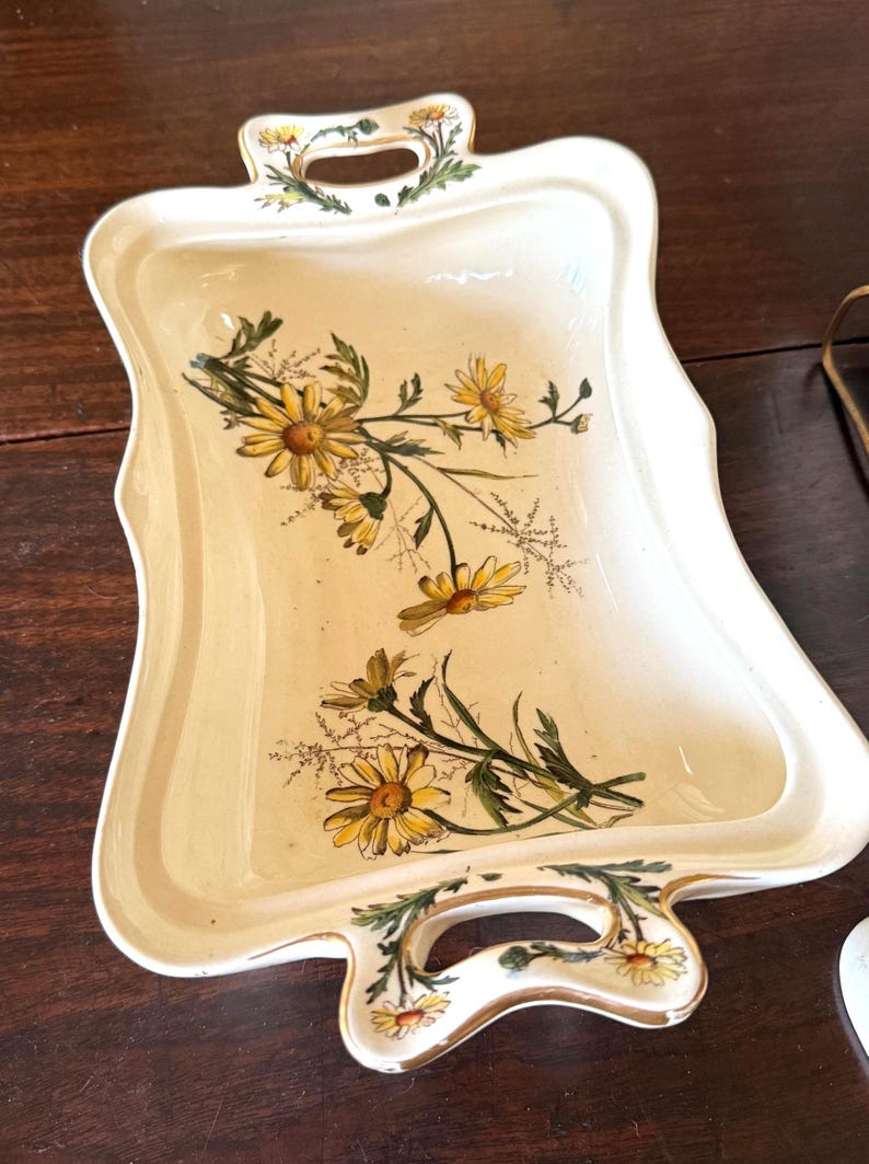May include: Cream-colored rectangular serving tray with a scalloped edge and two handles. The tray is decorated with yellow daisies and green leaves. The handles are adorned with floral accents. A decorative piece for serving.