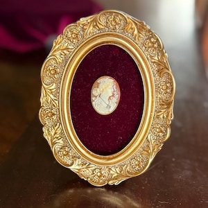 May include: Ornate gold-colored oval frame with a burgundy velvet backing. The frame surrounds a cameo portrait of a woman. The cameo is cream-colored with a detailed profile of a woman's face and neck. The frame has a detailed floral design.