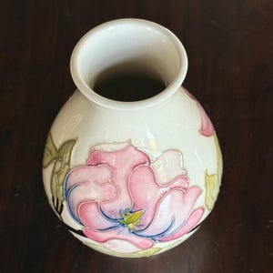 May include: Cream-colored ceramic vase with a large pink and blue floral design. The vase has a round body and a wide opening. The flower has green leaves and a yellow center. The vase is likely for decorative purposes.
