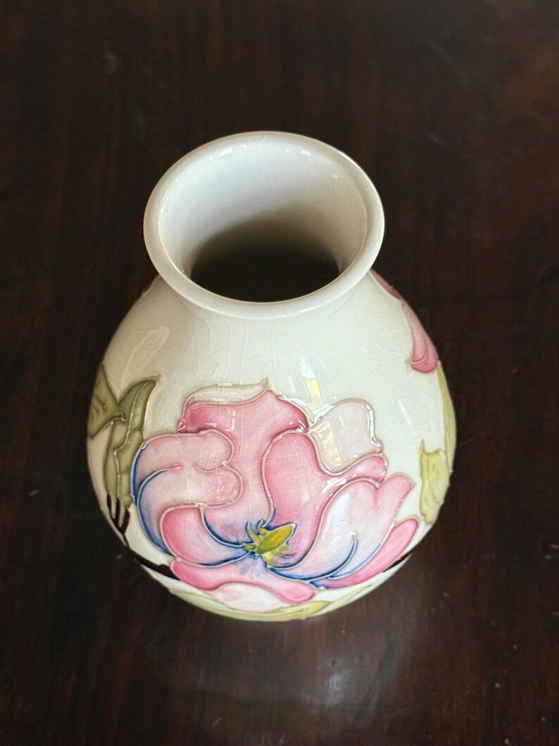May include: Cream-colored ceramic vase with a large pink and blue floral design. The vase has a round body and a wide opening. The flower has green leaves and a yellow center. The vase is likely for decorative purposes.