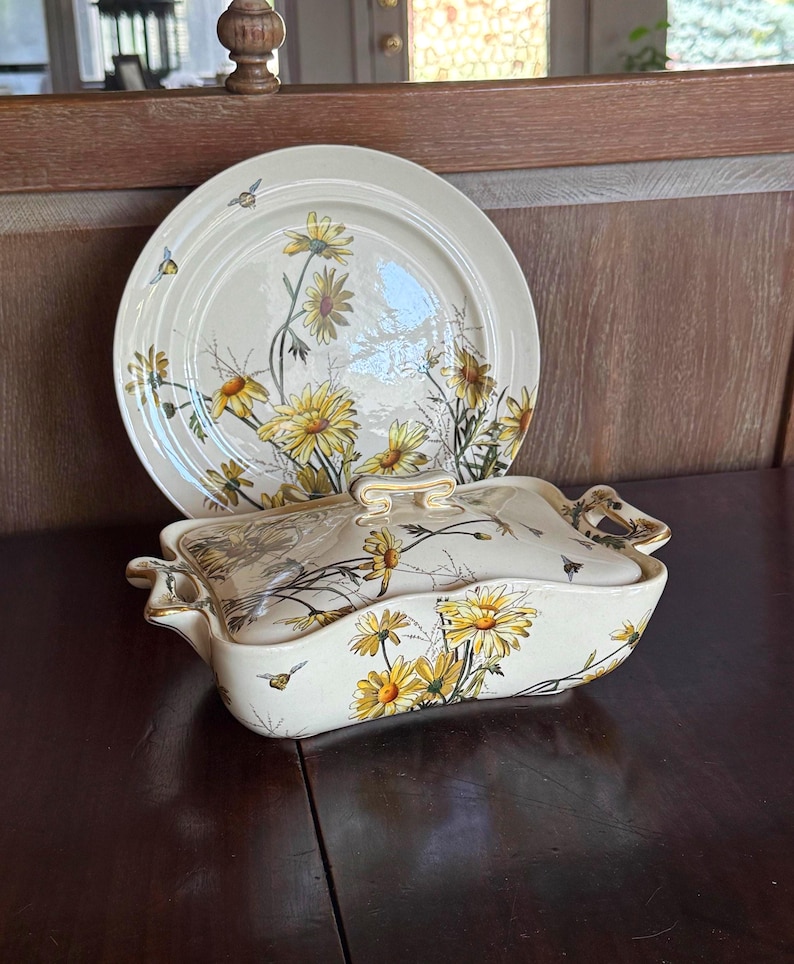 May include: Cream-colored ceramic serving dish and plate set. The set features a yellow daisy floral design with green stems and leaves, and small flying insects. The dish has a lid and handles, and the plate is round.
