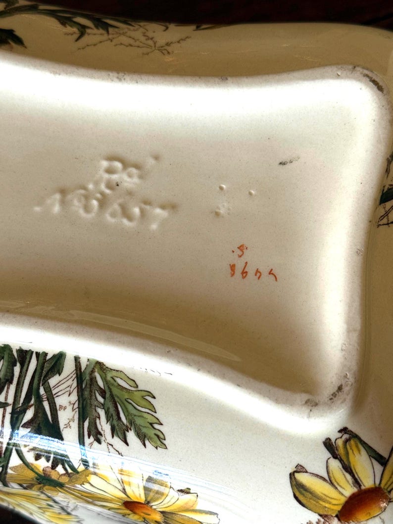 May include: Cream-colored ceramic serving dish with a floral design featuring yellow daisies and green leaves. The dish has a wavy shape and is marked with the numbers "18627" and "964".